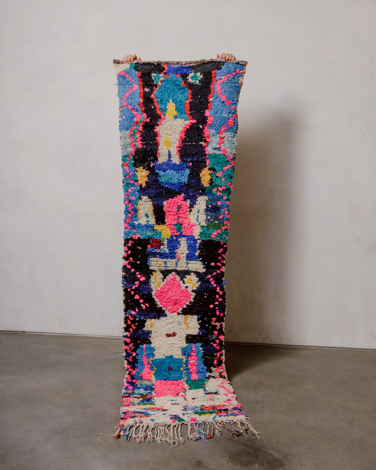 Berberlin boucherouite runner rug featuring blue, black and pink, being held up against a white wall.